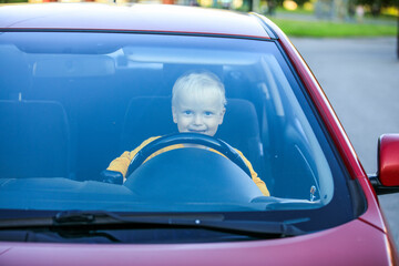 A little blond boy in a yellow jumper is driving a red car and smiling in a good mood. The green leaves of the trees are visible behind his back.