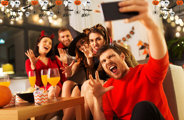 Poster - friendship, holiday and people concept - group of happy smiling friends in halloween costumes of vampire, devil, witch and cheetah taking selfie by smartphone at home party at night