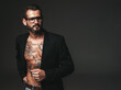 Portrait of handsome confident stylish hipster lambersexual model. Sexy modern man dressed in black jacket. Naked torso with tattoos.Fashion male posing in studio on dark background. In spectacles
