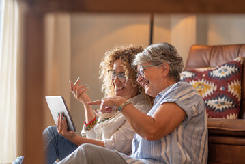 Adult beautiful daughter with elderly mother sitting at home looking social media on digital tablet