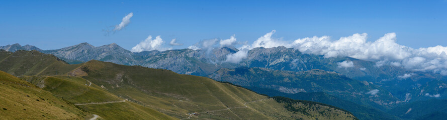 Wall Mural - Panoramic of the peaks of the mountain range between clouds in the background, above a clear blue sky, in the foreground the hill in front without trees, with grass, grass, for cattle, and a path that