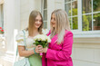 © MartaKlos - Young woman with blond hair in pink suit and smiling woman or girl with bouquet of flowers in hand happily looking at each other on wedding ceremony