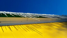 Dangerous Icicles. Sharp Icicles Hang From The Roof Of A Yellow Building Covered In Snow Against A Blue Sky. Winter Threats