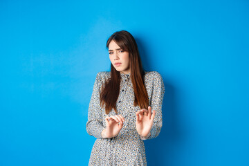 Disgusted and reluctant woman asking to keep away from here, showing block gesture, raising hands to stop you, standing on blue background