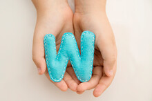 Stuffed Felt Letter Of The Alphabet In Open Hands Of Child. Kid Holding Handmade Capital Letter " W " In Isolated Milky White Background. Montessori Colors.