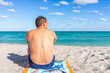 © Kristina Blokhin - Turquoise vibrant colorful water and back of tan man sitting on towel looking at sand shore in Sunny Isles Beach, North Miami, Florida during sunny day in winter