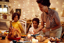 African American Mother Lights Candles During Thanksgiving Meal At Dining Table.