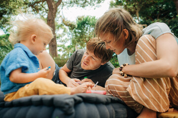 Wall Mural - Portrait of smiling couple painting with child outdoors. Mid adult parents resting on mattress with toddler daughter. Parenthood and creativity concept