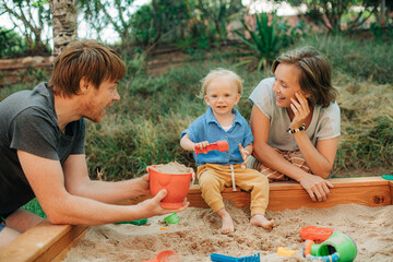 Wall Mural - Happy family with toddler child playing in sandbox. Toddler girl and her mother sitting at sandpit and father giving toy bucket with sand. Family outdoor activity concept