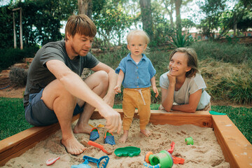 Wall Mural - Cheerful family playing together in sandpit. Portrait of mid adult parents sitting with their toddler daughter at sandbox. Family and childcare concept