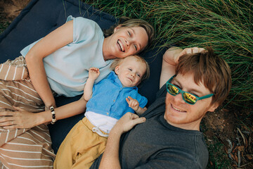 Wall Mural - Portrait of happy family resting on mattress. Cheerful parents with little child lying together on grass outdoors. Family vacation concept