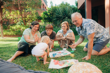 Wall Mural - Happy multi generation family playing with child outdoors. Parents and grandparents sitting on grass and painting together with toddler. Family leisure and creativity concept