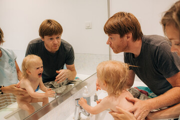 Wall Mural - Little girl crying while her parents washing her. Reflection of family with toddler in bathroom mirror. Childcare and hygiene concept