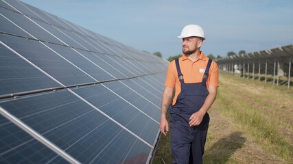 Wall Mural - Male engineer in uniform walking and looking at solar power plant. Man in hard helmet examining object. Concept of solar station development and green energy. Worker on solar power station outdoor