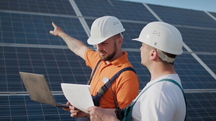 Wall Mural - Close up engineers use laptop and project plan on the solar farm in special uniform standing look around discuss the installation of sunny batteries electricity environmental. Green energy jobs