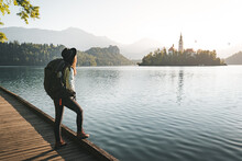 Hipester Young Woman With Backpack Enjoying Breathtaking  Sunrise By The Lake. Woman Wearing Hat And Denim Jacket Looking At View In Bled, Slovenia. Travel Freedom Lifestyle