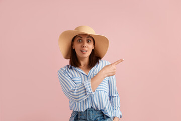 Wow, look here. Inspired woman points out at blank copy space with index finger, has eyes and mouth open, wears straw hat, striped shirt, jeans with belt, on pink background. Summer emotions concept.