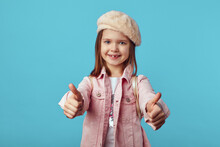 Charming Little Kid In Pink Jacket And Hat Smiling And Showing Thumbs Up Gesture Against Blue Studio Background