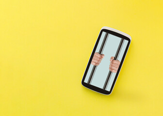 Creative, abstract idea of losing freedom inside the virtual world. Male hands in mobile phone isolated on illuminated yellow background hold prison bars. Minimalist and surrealist note card concept. 