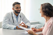 © insta_photos - Happy Indian male doctor consulting senior old patient filling form at consultation. Friendly professional physician talking to mature woman signing medical paper during appointment visit in clinic.
