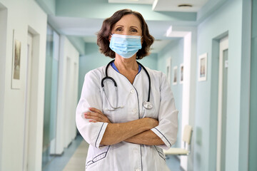 Wall Mural - Older female doctor wearing face mask and white medical coat standing in hospital hall. Senior mature woman professional physician in facemask looking at camera. Portrait