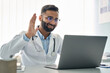 © insta_photos - Happy Indian male doctor waving hand having virtual video call online consultation on laptop computer in hospital. Tele health medicine. Telemedicine patients appointment distance remote chat in India