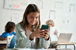 © insta_photos - Happy latin hispanic kid girl school student using smartphone in classroom. Preteen child holding mobile cell phone having fun with apps playing games and checking social media at school during break.