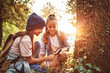 Two little girls in warm hats with backpacks examining tree bark through magnifying glass in forest