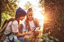 Two Little Girls In Warm Hats With Backpacks Examining Tree Bark Through Magnifying Glass In Forest