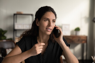 Millennial Latin business woman engaged in telephone talk, making call, speaking on mobile phone at work desk in home office, consulting customer support service, looking away, listening