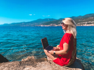 Attractive stylish blonde smiling young woman working typing on laptop. Freelancer on the beach in Opatija, Croatia. Girl with notebook at the sea, ocean. Freelance, online education concept