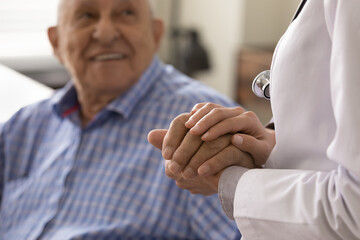 female therapist in white coat with stethoscope holding hand of optimistic senior patient, giving co