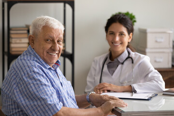 Wall Mural - Portrait of happy elder 80s man and young female doctor at appointment. Senior patient visiting therapist in office, looking at camera, smiling, Geriatric healthcare, elderly medic care concept