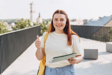 Wall Mural - Attractive young female tourist is exploring new city. Happy redhead Woman with map in hands showing thumb up gesture