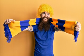 Wall Mural - Redhead man with long beard football hooligan cheering game wearing funny wig smiling looking to the side and staring away thinking.