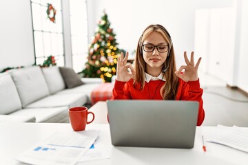 Poster - Young caucasian girl sitting on the table working using laptop by christmas tree relax and smiling with eyes closed doing meditation gesture with fingers. yoga concept.