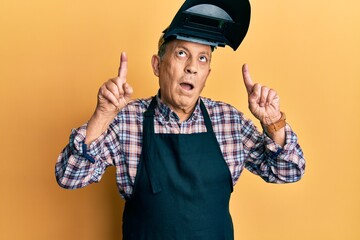 Poster - Handsome senior man with grey hair wearing welding protection mask amazed and surprised looking up and pointing with fingers and raised arms.