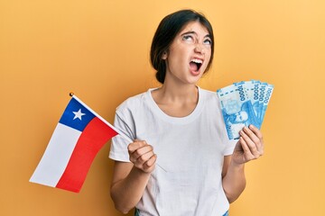 Wall Mural - Young caucasian woman holding chile flag and chilean pesos banknotes angry and mad screaming frustrated and furious, shouting with anger looking up.