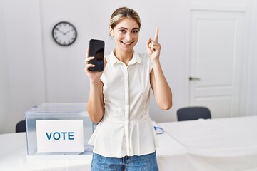 Canvas Print - Young blonde woman at political election holding smartphone smiling with an idea or question pointing finger with happy face, number one