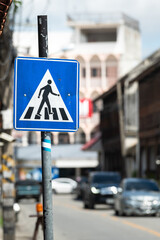 A pedestrian crosswalk signpost symbol in blue and triangle logo on the street with city as blurred background. Transportation sign object photo.