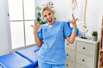 Canvas Print - Beautiful blonde physiotherapist woman working at pain recovery clinic shouting with crazy expression doing rock symbol with hands up. music star. heavy concept.