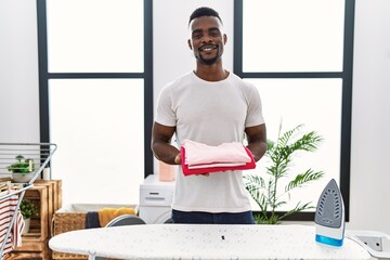 Sticker - Young african man holding folded laundry after ironing smiling with a happy and cool smile on face. showing teeth.