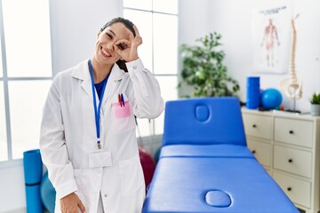 Canvas Print - Young doctor woman working at pain recovery clinic doing ok gesture with hand smiling, eye looking through fingers with happy face.