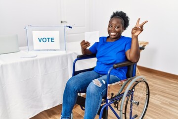 Sticker - Young african woman sitting on wheelchair voting putting envelop in ballot box smiling looking to the camera showing fingers doing victory sign. number two.