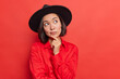 © WHstudio Leushin N - Stylish beautiful Asian woman keeps handon chin being deep in thoughts considers something wears fashionable black hatt shirt poses against red background blank copy space for advertisement.
