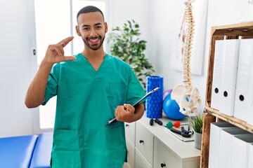 Poster - African american physiotherapist man working at pain recovery clinic smiling and confident gesturing with hand doing small size sign with fingers looking and the camera. measure concept.