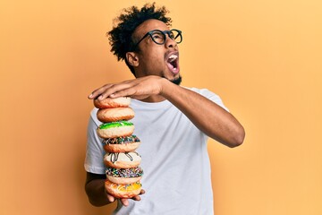 Young african american man with beard holding pile of tasty colorful doughnuts angry and mad screaming frustrated and furious, shouting with anger looking up.