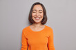 © wayhome.studio  - Sincere beautiful Asian woman glad to hear heartwarming words keeps eyes closed smiles gently has natural dark hair healthy skin dressed in casual orange jumper poses against grey background