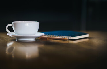 Focus on white coffe cup with reflection on wooden table with blue note book and pen, Dark tone