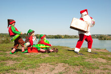 SANTA KLAUS AND ELF On The River Bank With A Suitcase In His Hands Carries Elves On Sleds In Inflatable Circles In The Summer For The New Year And Christmas